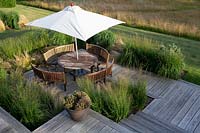 Meadow garden in Aldeburgh, Suffolk. High view of decked patio area with wooden table with parasol and chairs. Herbaceous border planting includes: Panicum virgatum - Heavy Metal, Molinia caerulea subsp. arundinacea - Transparent, Stipa tenuissima, Hakonechloa macra.