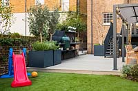 Modern garden with play area, outdoor kitchen with BBQ and dining area with big cubic containers with Olea europaea - Olive trees, Salvia officinalis and Betula pendula multi-stem 
