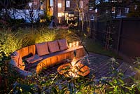 Seating area with fire pit and round wooden bench surrounded by Miscanthus sinensis 'Morning Light' - eulalia and Cornus alba 'Sibirica' Siberian dogwood AGM at night 