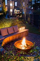 Seating area with fire pit and round wooden bench surrounded by Miscanthus sinensis 'Morning Light' -Eulalia and Cornus alba 'Sibirica' - Siberian dogwood, AGM at night in Autumn.