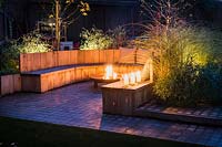 Seating area with fire pit and round wooden bench surrounded by Miscanthus sinensis 'Morning Light' - eulalia and Cornus alba 'Sibirica' - Siberian dogwood, AGM at night in Autumn.