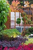 View across a garden planted in drifts of plants to a pavilion style Chinese garden, moongate, printed glass panels and a cloud pruned tree in a pot.