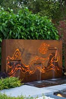 Freestanding rusty coreten steel fountain, featuring internally lit laser cut patterns reresenting mountains and clouds, with three rills behind pale grey stepping stones with a screen planting of Magnolias.