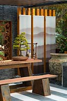 Corner detail of a pavilion with three glass screens, a decorative window, a large rustic pot, a timber slab bench with steel legs, a bonsai conifer tree.