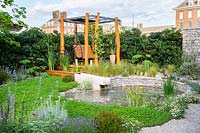 A garden with swing by the pond with water feature surrounded by ground covering plants. The Harmonious Garden of Life. RHS Chelsea Flower Show 2019