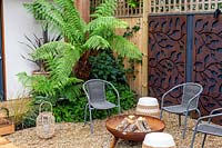 Contemporary garden in Wandsworth. With gravel patio, and chairs around a fire pit. In background Dicksonia antarctica  - tree fern, with decorative rusty metal panels on wooden fence with trellis