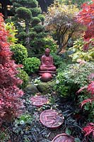 Red buddha statue set amongst autumnal colours and stepping stones at Four Seasons garden, Walsall, West Midlands, in October