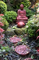 Red buddha statue set amongst beds autumnal colours and stepping stones at Four Seasons garden, Walsall, West Midlands.