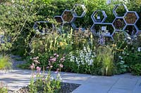Honeycomb shapes wall with twigs for solitary bees, surrounded by nectar-rich flowers  such as Campanula persicifolia var. alba, Buddleia, Rosa 'Kew Gardens', Achillea 'Terracotta', Verbascum 'Gainsborough', Daucus carota - The Urban Pollinator Garden - RHS Hampton Court Palace Garden Festival, 2019.