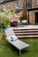 London contemporary garden patio with wooden steps leading up to modern garden sofa, chairs and table on patio with cedar batten trellis fencing behind. In foreground a modern sun lounger sits on artificial lawn.