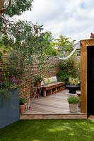 London contemporary garden - grey raised border on patio. Mixed summer planting. In background a lower wood deck area with Betula utilis and wooden seat. To the right a garden room sits behind an artificial lawn