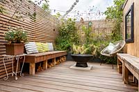 London contemporary garden -  lower wood deck area with  wooden seat and cushions. In the centre of the deck is a fire pit in front of a small raised border with Stipa tenuissima, Geranium johnsons blue, Magnolia grandiflora. To the right is a garden room or gym