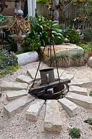 A firepit with a metal tripod and old copper pot with semi dressed sandstone blocks laid out in a sune pattern, mulched with seashells in front of sa garden with a sandstone boulder and a garden with succulents and Agapanthus.