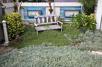 Shabby chic timber garden bench in front of a fence with recycled corrugated iron frames attached to a timber fence to display nautical themed objects, the bench is in a garden planted with Thyme and Gazanias as groundcovers.