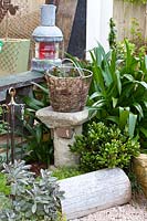 Detail of a garden with succulents and Agapanthus with a collection of objects featuring a timber log seat a stone plinth with a wire basket full of garden twine and a vintage railway lantern.