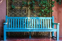 Turquoise bench in a shady corner with sunlight filtering through. Behind it a red plastered wall and ornately decorated metalwork gates.  Le Jardin Majorelle, Majorelle Garden, Marrakech