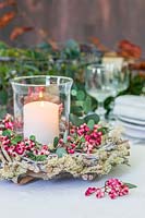 Table decoration comprised of: pillar candle in storm lantern surrounded by wooden wreath with moss, Sorbus - Rowan - berries and Eucalyptus foliage