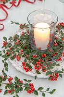 Pillar candle in storm lantern surrounded by white wicker matt and Cotoneaster berries and foliage.