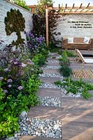 View along path made of paving and cobbles to pergola and bench. To one side decorative moss in shape of a tree on the stone wall next to the deck area. The 'Wetland Plants - The idea of Wilderness' garden 
