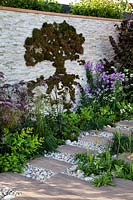 Decorative moss in shape of a tree on the stone wall in 'Wetland Plants - The idea of Wilderness' garden, flower bed and path of paving and cobbles nearby