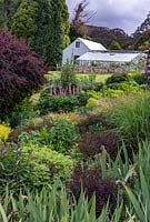 View across a herbaceous perennial border near shrubs to a traditional nursery glasshouse and a shed