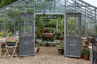 Traditional nursery glasshouse with double doors open for ventilation, showing a collection of potted plants, outdoor tables and chairs, the ground is mulched with pebbles