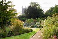 Gravel path in the front garden between grass and borders at the Old Vicarage, Weare, Somerset in August, with tower of Church of St Gregory beyond.