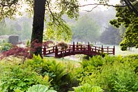 Red Nikko bridge in the Japanese garden in May at Heale Garden, Wiltshire with streams fringed with astilbes, Osmunda regalis, Matteuccia struthiopteris and acers in the foreground.