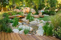 A natural looking stream and waterfall constructed from large boulders and small rocks, next to a small timber deck, interplanted with a variety of flowering and strappy leaved plants, featuring a low arched timber bridge and a small lawn area with circular seagrass cushions.
