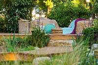 A timber deck with cushions, throws and seagrass seat, surroundings interplanted with a variety of shrubs, flowering and strappy leaved plants