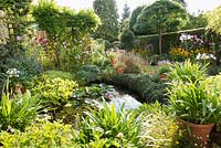 Pond in the back garden of a terraced house in Bristol in September, surrounded by lush planting including agapanthus, cotoneaster, cosmos, Erigeron karvinskianus, agastache and juniper
