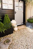 Deluxe garden shed of painted timber with door framed by standard olive, ceramic jars, clipped box with an ammonite inset into gravel in the foreground.