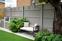 The barbeque grill and the bench with white cushions surrounded by Hydrangea and ferns by grey fence.