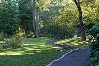 View from path of meandering, stone-lined stream running through landscape, with  green Adirondack chair set beneath trees