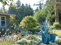 A modest front border includes a small patio, dramatic glass sculpture and fountain. Containers, glass art and plants are in shades of blue and white with purple and chartreuse accents