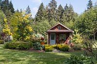 View across lawn to a rustic cabin set in a mixed bed of conifers, trees, shrubs and perennials