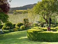 A lawn with low circular Buxus - Box - hedge around a vintage water pump and a mixed border, view of the apple orchards and hills beyond the garden boundary
