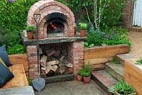 Outdoor brick oven with wooden seating and stone steps leading down from gravel path - RHS Malvern Spring Festival 