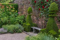 A bench flanked by box topiary, in a small formal, space planted with herbs, annuals and roses. The Herb Garden at Arley Hall, Cheshire, UK.

