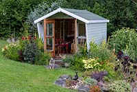 A summerhouse at the bottom of a hill, and edged in beds of daylilies, verbascums, dahlias and hostas. Bernie the schnoodle stands outside.