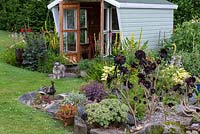 A summerhouse tucked away at the bottom of a hill, and edged in beds of daylilies, verbascums, dahlias, aeoniums and hostas. Bernie the schnoodle stands outside.