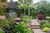 Irregular, sixth-of-an-acre town plot has a thatched  summerhouse and deck overlooking a bog garden of astilbes, arum lilies, hostas, ligularia and rheum. Pond crossed by a bridge leading to mirror on back boundary. Trees: silver birch, weeping pear and Japanese maple.