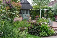 A thatched  summerhouse and deck overlook a bog garden of astilbes, arum lilies, hostas, ligularia and rheum. A bridge leads to mirror on back boundary, passing a silver birch.