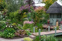 Sixth-of-an-acre, irregular plot has an arch clad in Rosa 'Veilchenblau' framing view of herbaceous borders, and flanked by acers. A thatched  summerhouse and deck overlook a pond and bog garden with arum lilies, rheum, ligularias and astilbes.