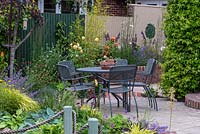 Sheltered dining area on terrace, beside curving, brick edged raised bed planted with roses, salvias, catmint and Verbena bonariensis.