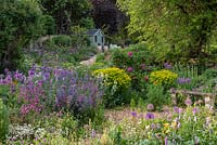 A stone bench sits in a small gravel garden edged in alliums, bistort, Euphorbia palustris, ragged robin, roses and catmint. Beyond, a paved path ascends to a summerhouse, passing a border in which flag irises and nectaroscordum star.