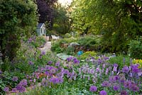 View through Allium 'Purple Sensation', Persicaria bistorta 'Superba', euphorbia, ragged robin, orlaya and catmint, to a paved path that ascends to a summerhouse, passing a border in which flag irises and nectaroscordum star. Beyond stands an Acer platanoides 'Drummondii'.