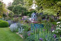 A gravel bed is planted with silver lamb's ears, alliums, irises and centranthus, surrounding a large metal drum of red dahlias and ivy. Beyond, a summerhouse overlooks a terrace and herbaceous bed punctuated with rose obelisks.