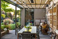 A covered dining area runs along the side wall of a contemporary courtyard. Reflections of the planting in the house windows.