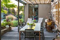 A covered dining area runs along the side wall of a contemporary courtyard. Reflections of the planting in the house windows.